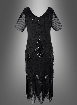 Das 20er Paillettenkleid mit transparenten Ärmeln „Gitta“ ist ein schwarzes, knielanges Kleid mit transparenten kurzen Ärmeln, eleganten Perlenverzierungen, funkelnden Pailletten-Details, die an ein klassisches Paillettenkleid für Damen erinnern, sowie verspielten Fransen am Saum, präsentiert vor einem grauen Hintergrund mit Farbverlauf.