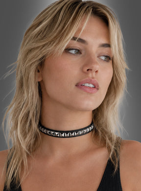 Eine Person mit blonden, welligen Haaren trägt ein ärmelloses schwarzes Oberteil und einen schwarzen Nieten-Rock im Rocker-Punk-Stil aus Kunstleder mit Pyramidennieten und blickt vor einem schlichten Hintergrund leicht zur Seite.