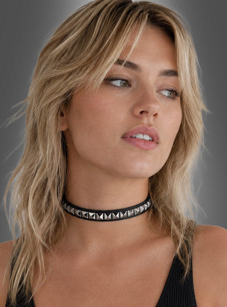 Eine Person mit blonden, welligen Haaren trägt ein ärmelloses schwarzes Oberteil und einen schwarzen Nieten-Rock im Rocker-Punk-Stil aus Kunstleder mit Pyramidennieten und blickt vor einem schlichten Hintergrund leicht zur Seite.