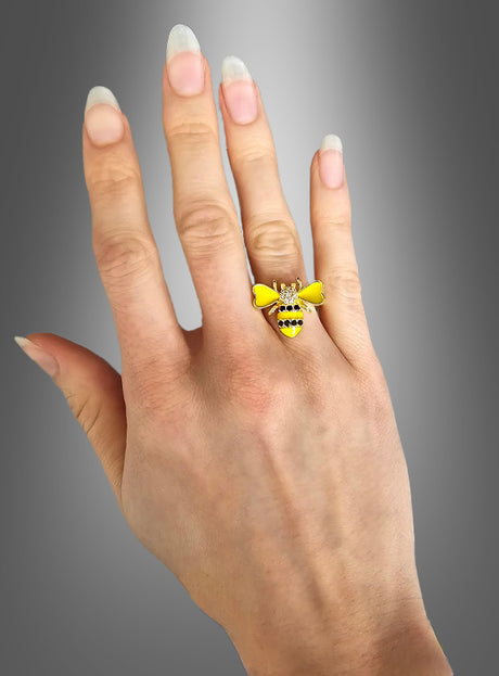 Eine Hand mit langen Naturnägeln trägt am Mittelfinger einen verstellbaren Bienenring mit Strasssteinen in Form einer Biene vor einem grauen Farbverlaufshintergrund – perfekt, um Ihrem Look eine verspielte Note mit Bienenkostüm-Schmuck zu verleihen.