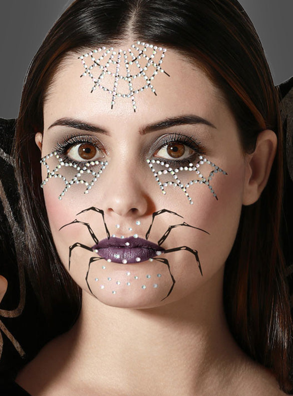 Eine Frau mit dunklem Haar präsentiert ein Halloween-Make-up, das von Spinnennetzen oberhalb und unterhalb ihrer Augen inspiriert ist und durch Spinnen-Gesichtstattoos hervorgehoben wird. Abgerundet wird der Look durch ein Spinnenmotiv um ihren Mund herum, violetten Lippenstift, selbstklebende Gesichtstattoos und funkelnde Strassakzente.