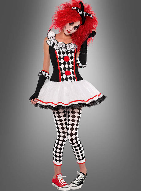 Eine Person, die das „Clownfrau Harlekin“-Kostüm trägt – bestehend aus einem schwarz-weiß-roten Harlekin-Tutu-Kleid, karierten Leggings, ungleichen Schuhen, einer leuchtend roten Perücke und auffälligem Make-up – steht vor einem grauen Hintergrund.