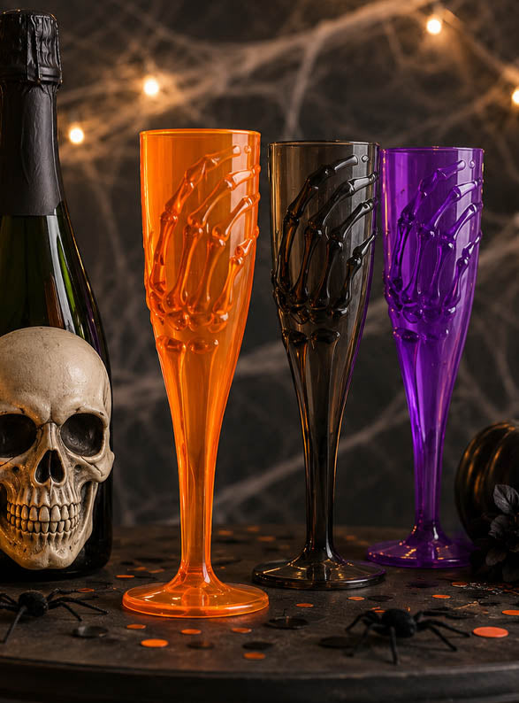 Drei „Skeletthand“-Sektgläser (20 cm) in Orange, Schwarz und Lila sind neben einem Totenkopf und einer Champagnerflasche auf einem Halloween-Tisch angeordnet, der mit Spinnen geschmückt und von Lichterketten beleuchtet ist – perfekt als gruselige Halloween-Tischdekoration für Ihre nächste Gothic-Party.