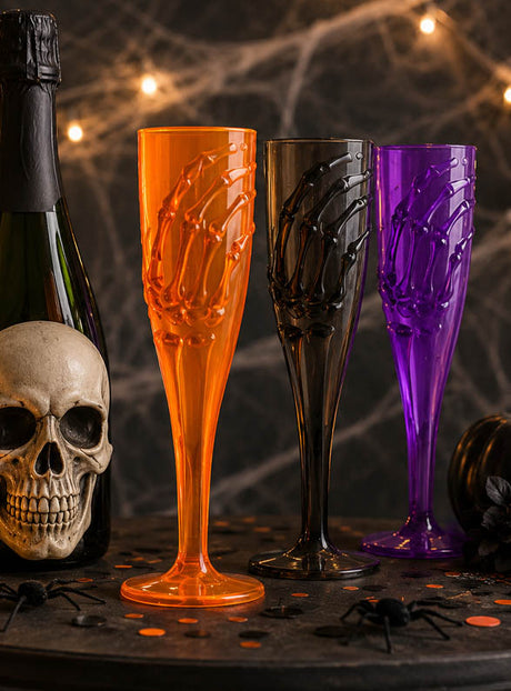 Drei „Skeletthand“-Sektgläser (20 cm) in Orange, Schwarz und Lila sind neben einem Totenkopf und einer Champagnerflasche auf einem Halloween-Tisch angeordnet, der mit Spinnen geschmückt und von Lichterketten beleuchtet ist – perfekt als gruselige Halloween-Tischdekoration für Ihre nächste Gothic-Party.