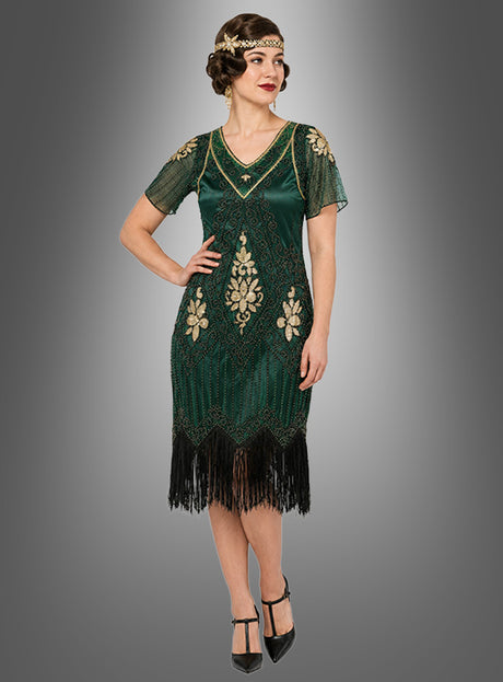 Eine Frau präsentiert das „20er Paillettenkleid mit transparenten Ärmeln Gitta“, ein grünes Flapperkleid im Stil der 1920er Jahreim Stil der 1920er Jahre, das mit goldenen Verzierungen und einem Fransenbesatz am Saum verziert ist, kombiniert mit schwarzen Pumps vor einem grauen Farbverlaufshintergrund – eine elegante Wahl für jede 20er-Jahre-Party oder als raffiniertes Paillettenkleid für Damen.