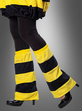 Eine Person, die schwarze Leggings, schwarze Schuhe mit hohen Absätzen und die „Biene Beinstulpen Plüsch schwarz-gelb“ als verspieltes Accessoire vor einem grauen Hintergrund trägt.