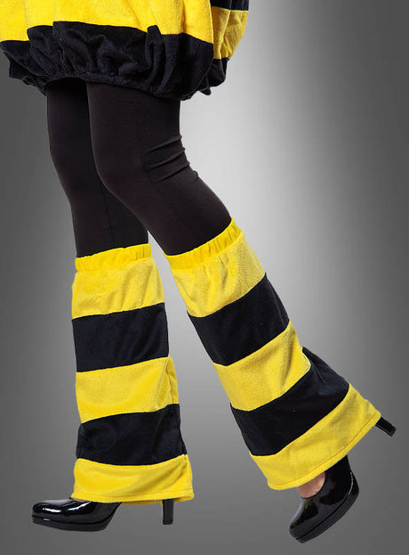 Eine Person, die schwarze Leggings, schwarze Schuhe mit hohen Absätzen und die „Biene Beinstulpen Plüsch schwarz-gelb“ als verspieltes Accessoire vor einem grauen Hintergrund trägt.