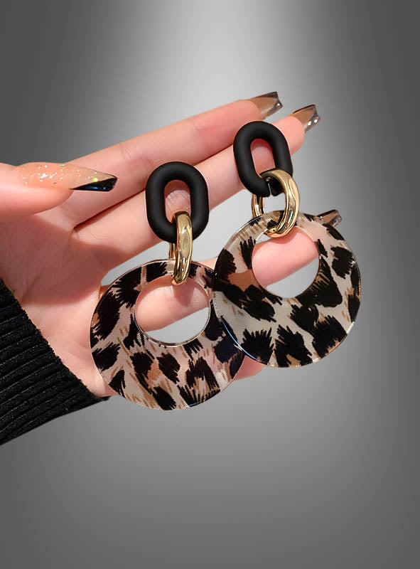 Eine Hand hält ein Paar „Leopard“-Ohrringe (Modeschmuck), die sich durch schwarze, goldene und mit Leopardenmuster verzierte runde Akzente auszeichnen – ideal für Liebhaber von auffälligem Statement-Schmuck – vor einem grauen Hintergrund mit Farbverlauf.