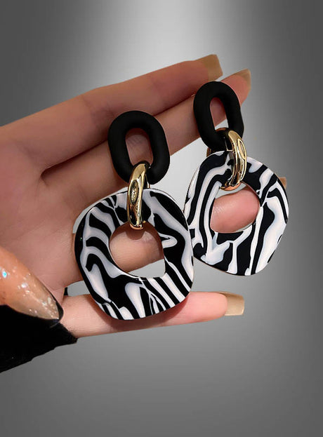 Eine Hand hält ein Paar „Zebra“-Ohrringe (Modeschmuck) – abstrakte Ohrhänger in Schwarz, Weiß und Gold mit marmoriertem Muster und geometrischen Formen, die von Accessoires der 80er Jahre inspiriert sind. Diese auffälligen Statement-Stücke werden vor einem grauen Hintergrund mit Farbverlauf präsentiert.