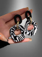 Eine Hand hält ein Paar „Zebra“-Ohrringe (Modeschmuck) – abstrakte Ohrhänger in Schwarz, Weiß und Gold mit marmoriertem Muster und geometrischen Formen, die von Accessoires der 80er Jahre inspiriert sind. Diese auffälligen Statement-Stücke werden vor einem grauen Hintergrund mit Farbverlauf präsentiert.
