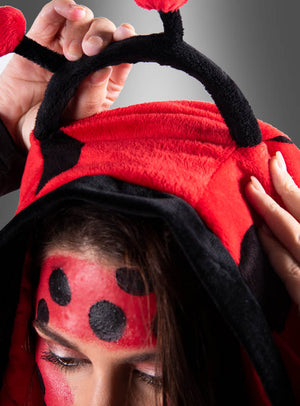 Person, die den „Marienkäfer-Poncho aus Plüsch“ in den Größen XS–XXXL trägt, mit roter Kapuze, schwarzen Fühlern und einer von Marienkäfern inspirierten Gesichtsbemalung, die die Kapuze mit beiden Händen hochhält – ideal als lustiges Karnevalskostüm oder als Teil eines verspielten Gruppenkostüms.