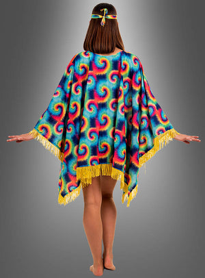 Eine Person steht barfuß mit ausgestreckten Armen da und trägt den „Hippie-Poncho für Damen mit Batik-Muster“ mit bunten Mustern und gelben Fransen sowie ein passendes Stirnband; die Aufnahme zeigt sie von hinten vor einem neutralen Hintergrund mit Farbverlauf.