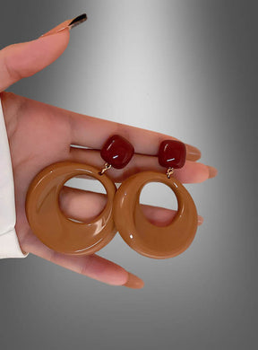 Eine Hand hält ein Paar braune Modeschmuck-Ohrringe im 70er-Jahre-Stil, bestehend aus großen braunen runden Creolen und roten quadratischen Ohrsteckern, vor einem grauen Hintergrund mit Farbverlauf, wodurch der markante Vintage-Stil von Retro-Acrylschmuck besonders gut zur Geltung kommt.