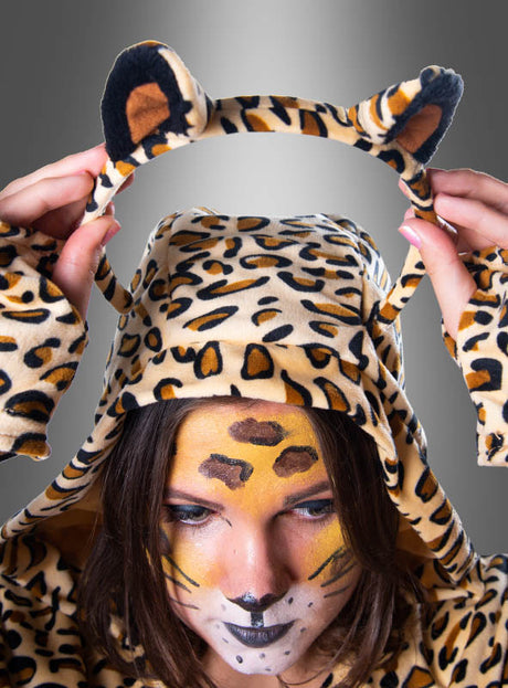 Eine Person, die einen Plüsch-Leoparden-Poncho in Größe XXL trägt, mit Gesichtsbemalung, die ein passendes Stirnband mit Katzenohren in der Hand hält und nach unten schaut.