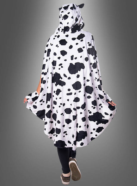 Eine Person steht mit dem Rücken zur Kamera vor einem grauen Hintergrund und trägt den „Kuh-Poncho aus Plüsch mit Eutertasche“ (Größen S–XXXL), ein kuscheliges Cape mit Kapuze im schwarz-weißen Kuhmuster.