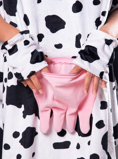 Eine Person, die den „Kuh-Poncho aus Plüsch mit Eutertasche“ in den Größen S–XXXL sowie fingerlose Handschuhe trägt und ihre Hände in die rosa, euterförmige Vordertasche des Plüsch-Outfits steckt.