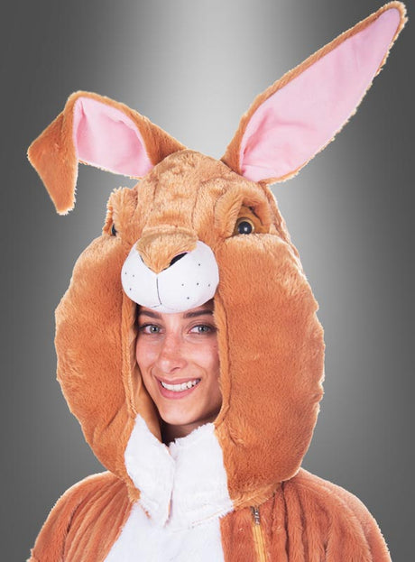 Eine Person lächelt, während sie das „Deluxe Osterhasen-Kostüm aus waschbarem Plüsch“ trägt – ein großes braun-weißes Hasenkostüm mit markanten Ohren und rosa Innenfutter –, vor einem grauen Hintergrund mit Farbverlauf.