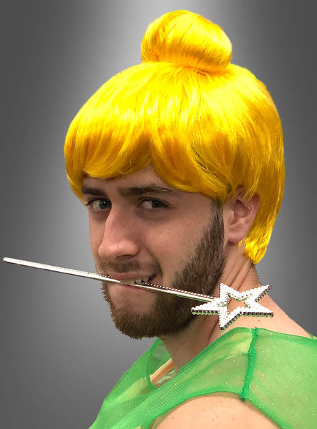 Eine Person, die eine „Gelbe Fee“-Perücke mit Dutt und ein grünes Outfit trägt, hält einen Zauberstab mit Sternspitze im Mund und blickt vor einem grauen Hintergrund in die Kamera.