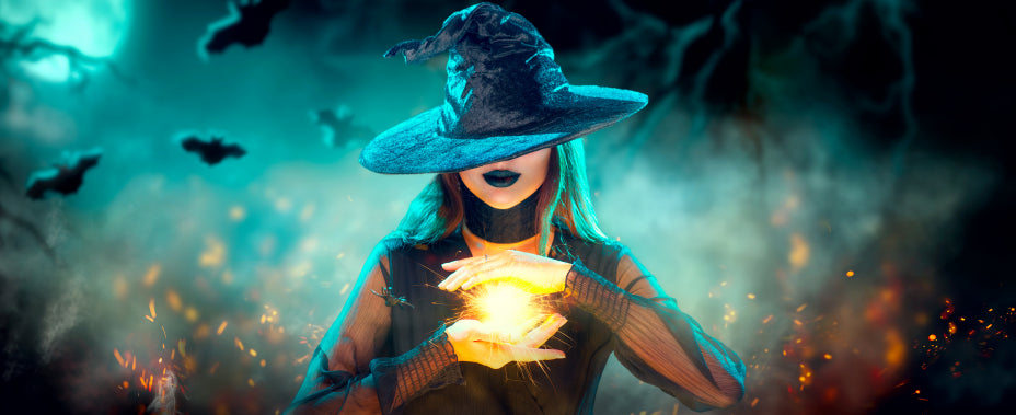 Eine Person in einem Hexenkostüm hält mit beiden Händen eine leuchtende Kugel in den Händen, umgeben von Nebel, Fledermäusen und einem leuchtenden Mond im Hintergrund.