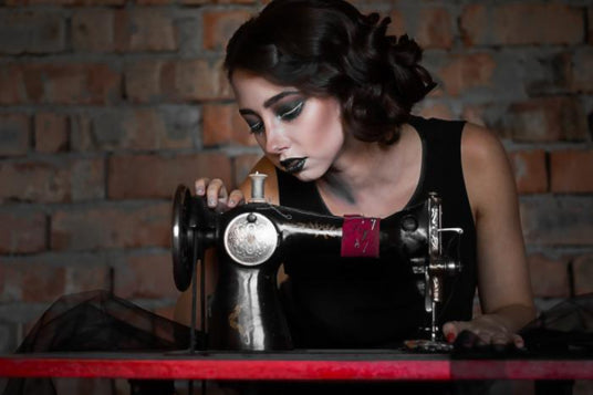 Eine Person mit auffälligem Make-up sitzt an einer altmodischen Nähmaschine, ist ganz in ihre Arbeit vertieft, im Hintergrund ist eine Backsteinmauer zu sehen.