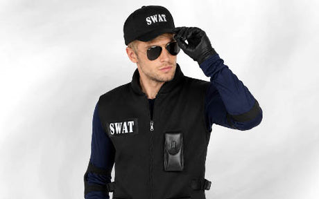 Eine Person, die eine SWAT-Weste, eine Mütze, schwarze Handschuhe und eine Sonnenbrille trägt, steht vor einem schlichten Hintergrund und hält die Sonnenbrille mit einer Hand fest.