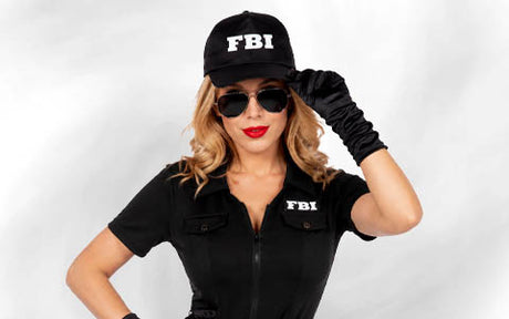 Eine Frau in einem schwarzen FBI-Kostüm, mit Sonnenbrille und Mütze steht vor einem hellen Hintergrund und berührt mit einer behandschuhten Hand den Rand ihrer Mütze.