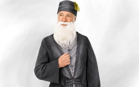 Ein älterer Mann mit weißem Bart, der eine dunkle traditionelle Robe und einen Hut mit gelber Quaste trägt und vor einem schlichten, hellen Hintergrund steht.