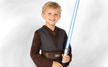 Ein lächelnder kleiner Junge in einem braunen Kostüm hält ein leuchtend blaues Lichtschwert in der Hand und steht vor einem schlichten weißen Hintergrund.
