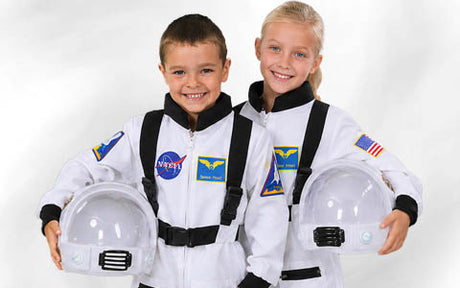 Zwei Kinder in weißen Astronautenanzügen mit NASA-Aufnähern, die lächeln und Weltraumhelme in die Höhe halten, vor einem schlichten weißen Hintergrund.