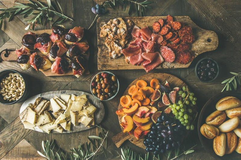 Eine Auswahl an Wurst- und Käsewaren, Käse, Brot, Obst, Nüssen und Oliven, angerichtet auf Holzbrettern und in Schalen auf einem rustikalen Tisch.