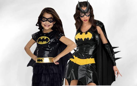 Zwei Personen in Batgirl-Kostümen stehen nebeneinander; sie tragen schwarze Masken, Umhänge und Outfits mit gelben Batman-Logos auf der Brust.