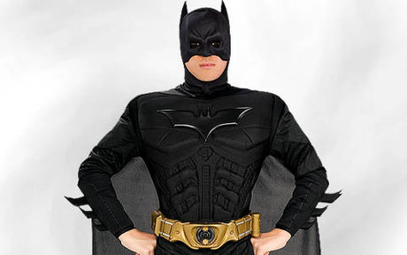 Eine Person in einem schwarzen Batman-Kostüm mit Umhang und goldenem Werkzeuggürtel, die mit den Händen in den Hüften vor einem schlichten, hellen Hintergrund steht.