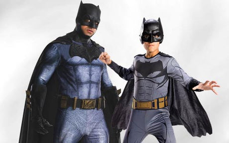 Zwei Personen in Batman-Kostümen posieren nebeneinander vor einem hellen Hintergrund – ein Erwachsener und ein Kind.