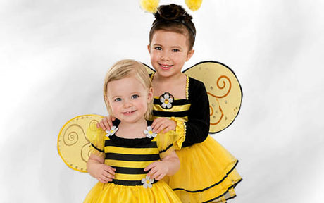 Zwei kleine Kinder, die identische gelb-schwarze Bienenkostüme mit Flügeln und Blumendekorationen tragen, stehen nebeneinander vor einem schlichten Hintergrund und lächeln in die Kamera.