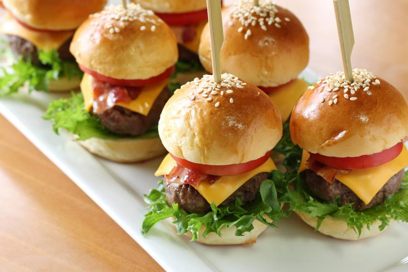 Eine Reihe von Mini-Cheeseburgern mit Salat, Tomate, Käse und Speck auf Sesambrötchen, die jeweils mit einem Holzspieß zusammengehalten werden und ordentlich auf einer weißen Platte angerichtet sind.