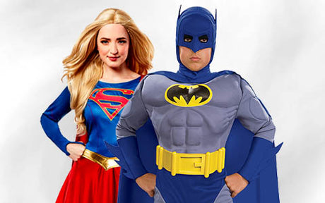 Eine Frau in einem Supergirl-Kostüm und ein Mann in einem Batman-Kostüm stehen nebeneinander vor einem einfarbigen Hintergrund.