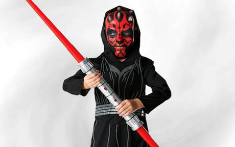 Ein Kind in einem schwarzen Kostüm mit einer rot-schwarzen Maske hält vor einem schlichten weißen Hintergrund ein rotes Spielzeug-Lichtschwert mit zwei Klingen in der Hand.