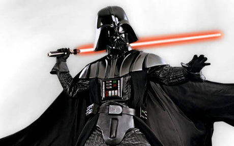 Vor einem schlichten weißen Hintergrund steht eine als Darth Vader verkleidete Person in schwarzer Rüstung und einem Umhang, die ein rotes Lichtschwert in einer erhobenen Hand hält.