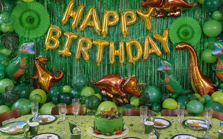 Dekoration für eine Geburtstagsparty zum Thema Dinosaurier mit grünen und goldenen Dekorationselementen, „Happy Birthday“-Luftballons, Dinosaurier-Luftballons und einer grünen Torte auf einem geschmückten Tisch.