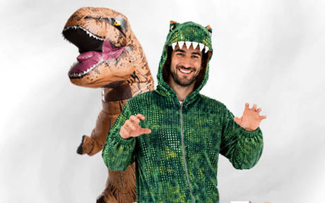 Ein Mann in einem grünen Dinosaurierkostüm posiert und lächelt, während hinter ihm vor einem schlichten Hintergrund eine Person in einem aufblasbaren T-Rex-Kostüm steht.