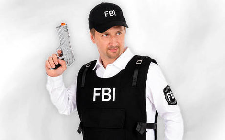 Ein Mann, der eine FBI-Weste und eine FBI-Kappe trägt, hält eine Spielzeugpistole in der Hand und steht vor einem schlichten, hellen Hintergrund.