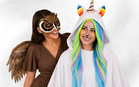 Zwei Frauen in Kostümen: Die eine trägt eine braune Eulenmaske und Flügel, die andere einen Einhorn-Strampler mit Regenbogenhaaren und einem Horn; beide lächeln.