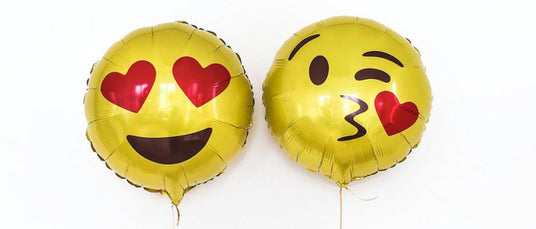 Zwei Goldfolienballons in Form von Emoji-Gesichtern; einer mit Herzaugen und einem Lächeln, der andere zwinkert und wirft einen Kuss mit einem roten Herzen zu.