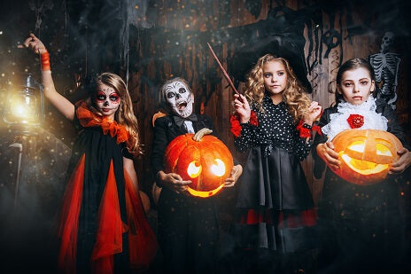 Vier Kinder in Halloween-Kostümen stehen drinnen, halten geschnitzte Kürbisse und eine Laterne in den Händen und sind von gruseligen Dekorationen und gedämpftem Licht umgeben.