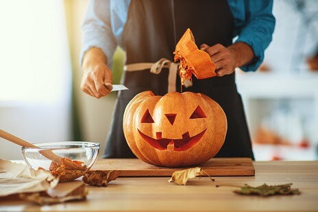 Eine Person in einer Schürze schnitzt auf einem Holztisch einen Kürbis zu einer Jack-o-Lantern, daneben liegen eine Schüssel, Blätter und Küchenutensilien.