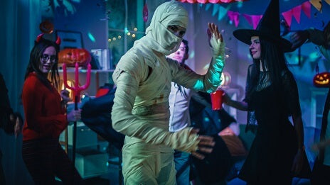Eine Gruppe von Menschen in Halloween-Kostümen, darunter eine Mumie, eine Hexe und ein Teufel, tanzt und feiert auf einer Party, die mit Lichtern und Kürbissen geschmückt ist.