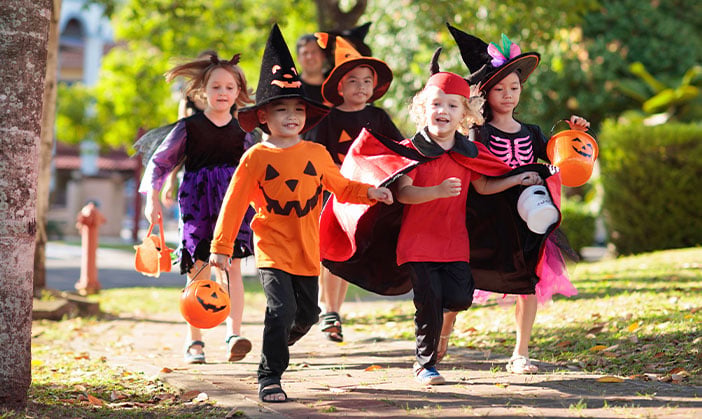 Eine Gruppe von Kindern in Halloween-Kostümen läuft an einem sonnigen Tag draußen spazieren, hält kürbisförmige Eimer in den Händen und lächelt.