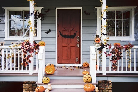 Die Veranda ist zu Halloween mit geschnitzten Kürbissen, Herbstblättern, Spinnweben und schwarzen Fledermaus-Ausschnitten geschmückt, die neben einer roten Tür hängen.
