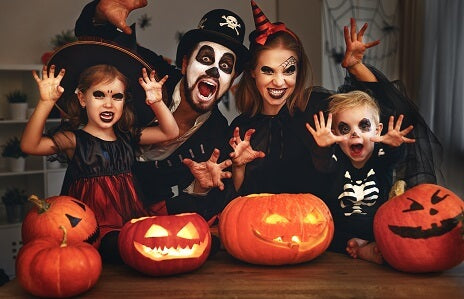 Eine Familie in Halloween-Kostümen posiert mit geschnitzten Kürbislaternen auf einem Tisch und schneidet dabei gruselige Grimassen in die Kamera.