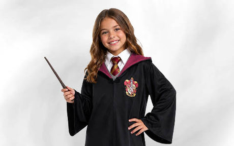 Ein junges Mädchen in einer schwarzen Robe mit Wappen, das einen Zauberstab in der Hand hält und lächelt, in einem von Hogwarts inspirierten Kostüm vor einem einfarbigen Hintergrund.